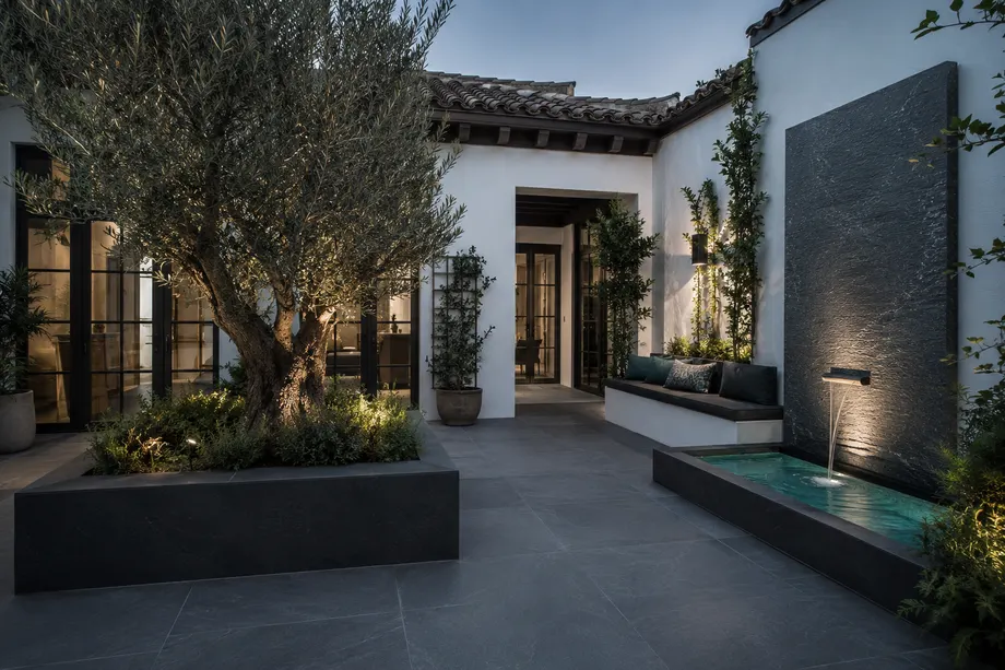 Patio renovado con pavimento porcelánico grafito, olivo en jardinera elevada y fuente de pared